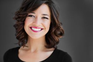 Professional headshot of someone with dark wavy hair and bright smile against dark background.