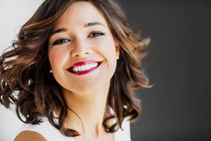 Professional portrait showcasing wavy brown hair, bright smile and natural makeup against dark background.