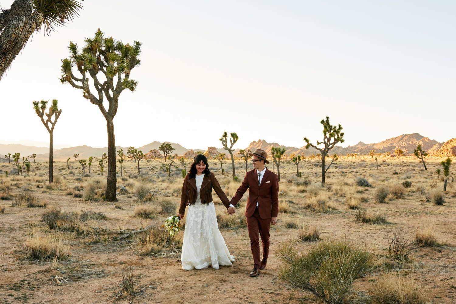 Two figures in wedding attire walk hand in hand through a desert landscape with iconic Joshua Trees in Joshua Tree National Park.