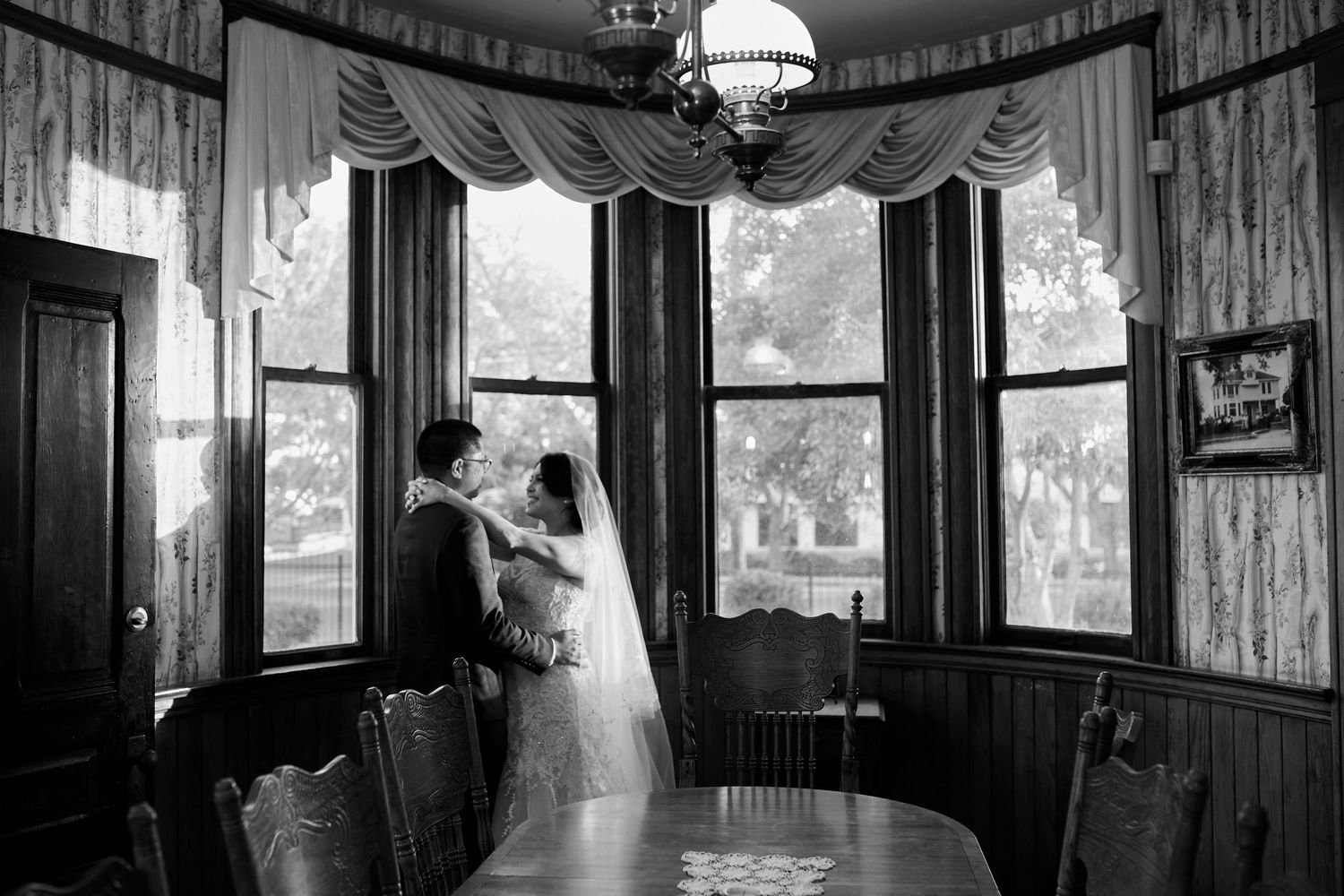 Two people converse in an elegant Victorian-style bay window dining room in black and white.