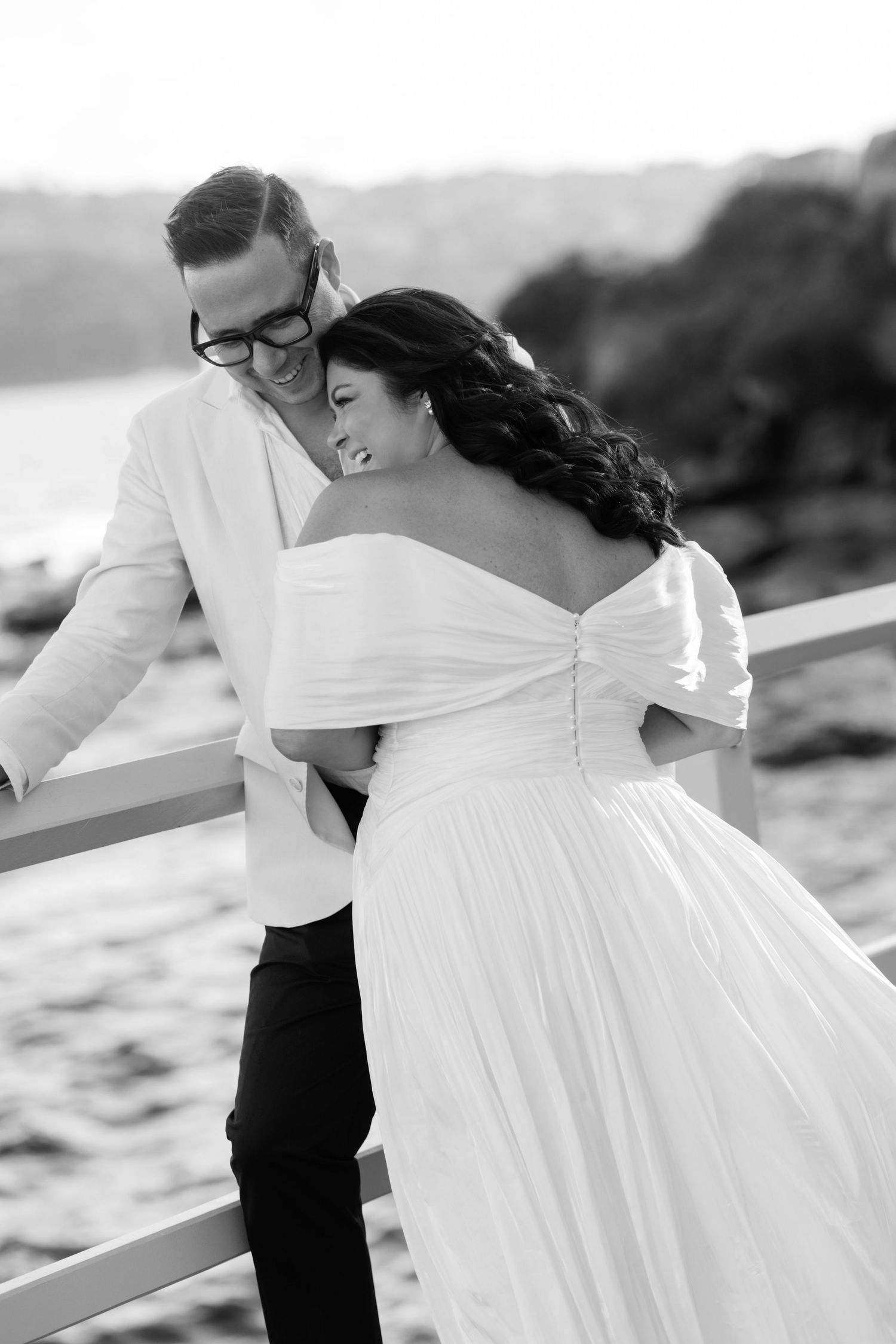 A couple in formal wedding attire embrace on a road with a hillside and trees in the background at sunset.