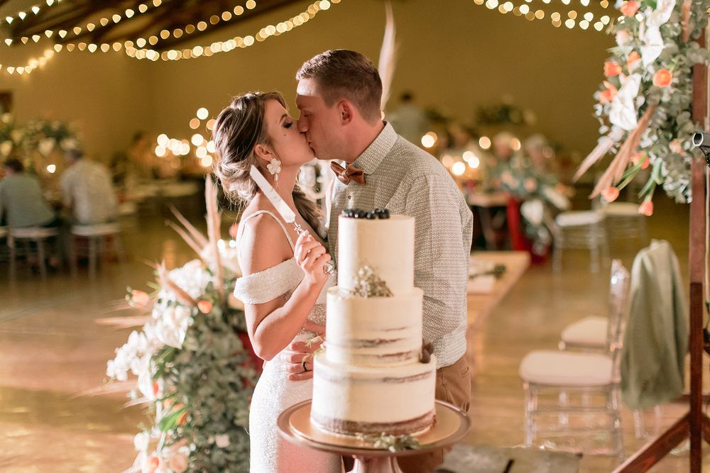 Bride and groom sharing a kiss behind a white wedding cake at a romantic reception with fairy lights.