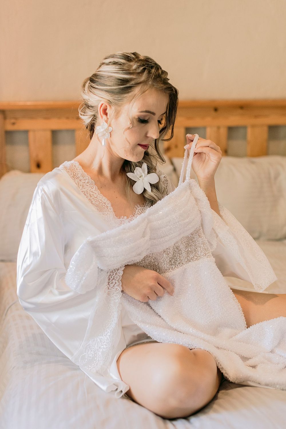 Bride in white lace robe sitting on bed holding lingerie before wedding preparations.