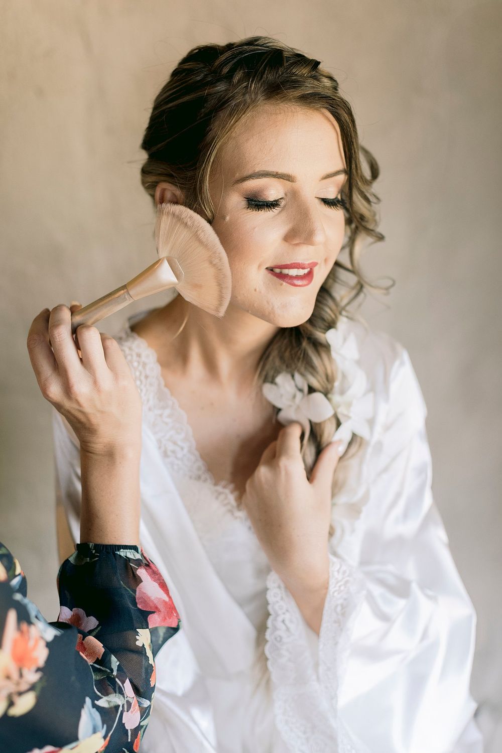 Bride in white lace robe with eyes closed as makeup artist applies powder brush to face.