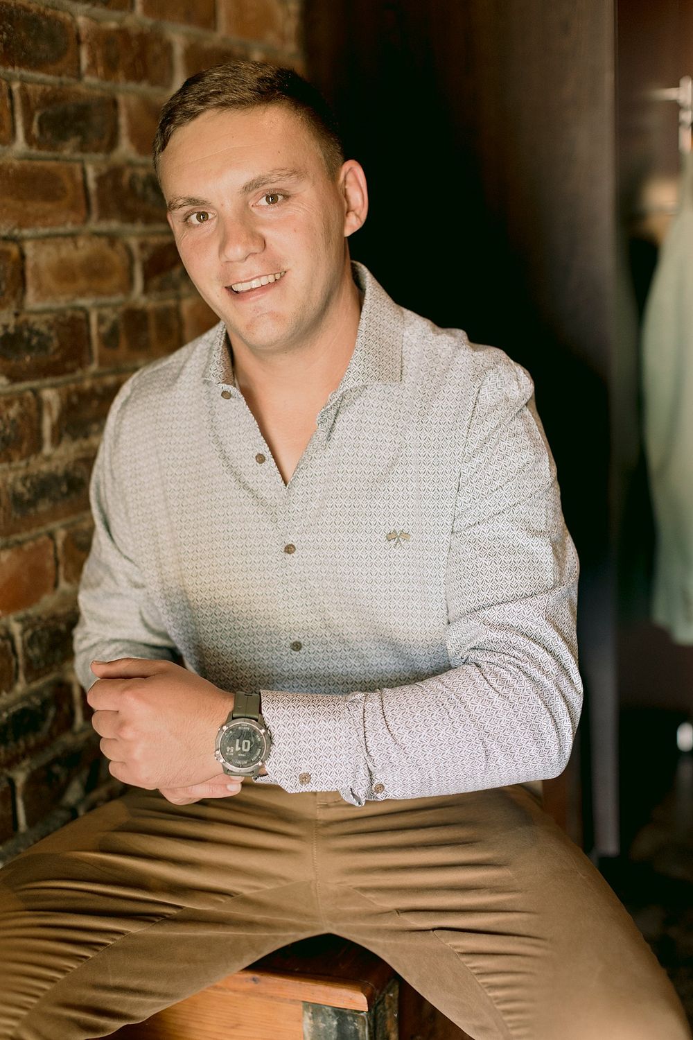 Smiling groom in grey patterned shirt and khaki pants sitting against brick wall background.