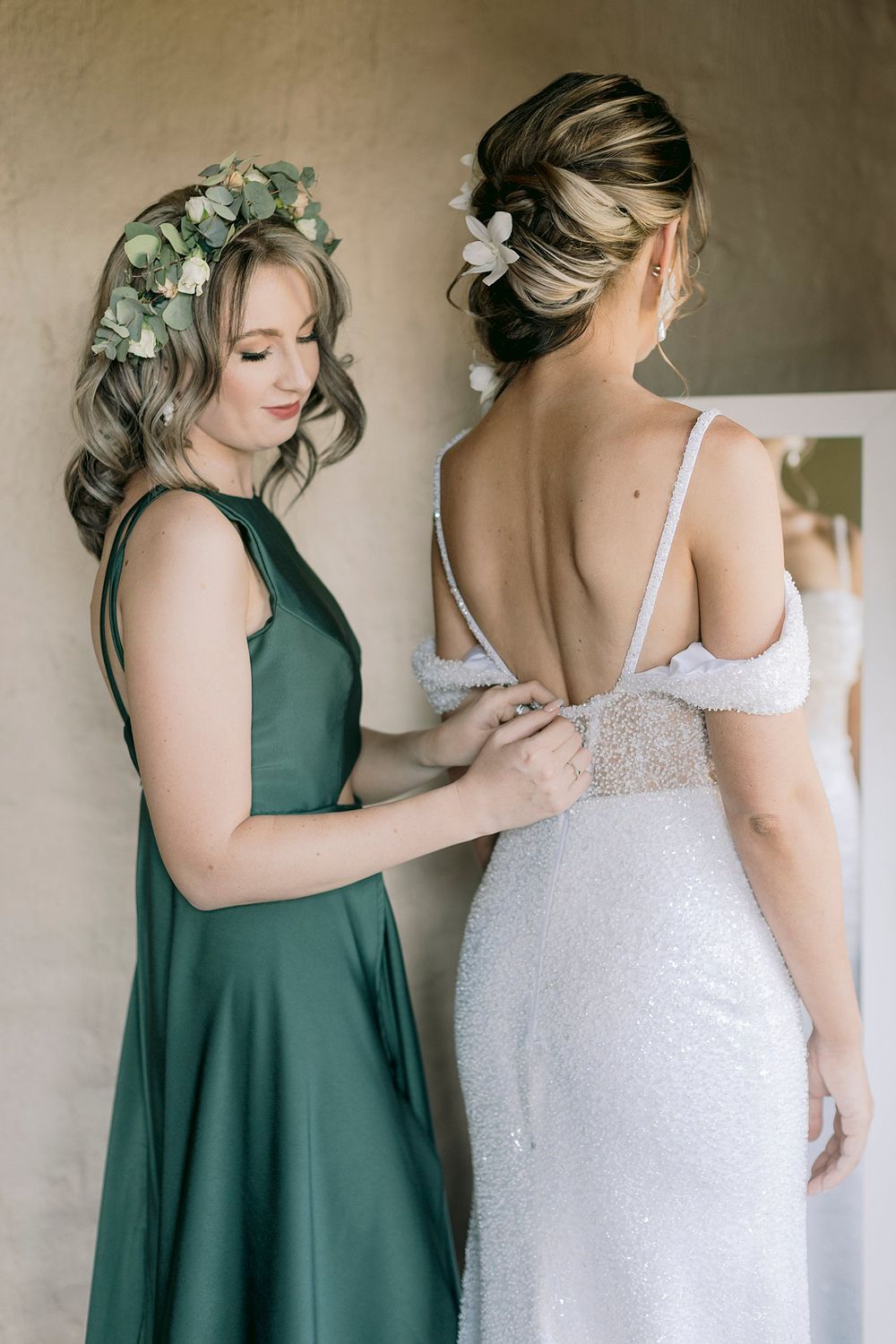Bridesmaid in green dress fastens the back of a sparkly white wedding gown for the bride.