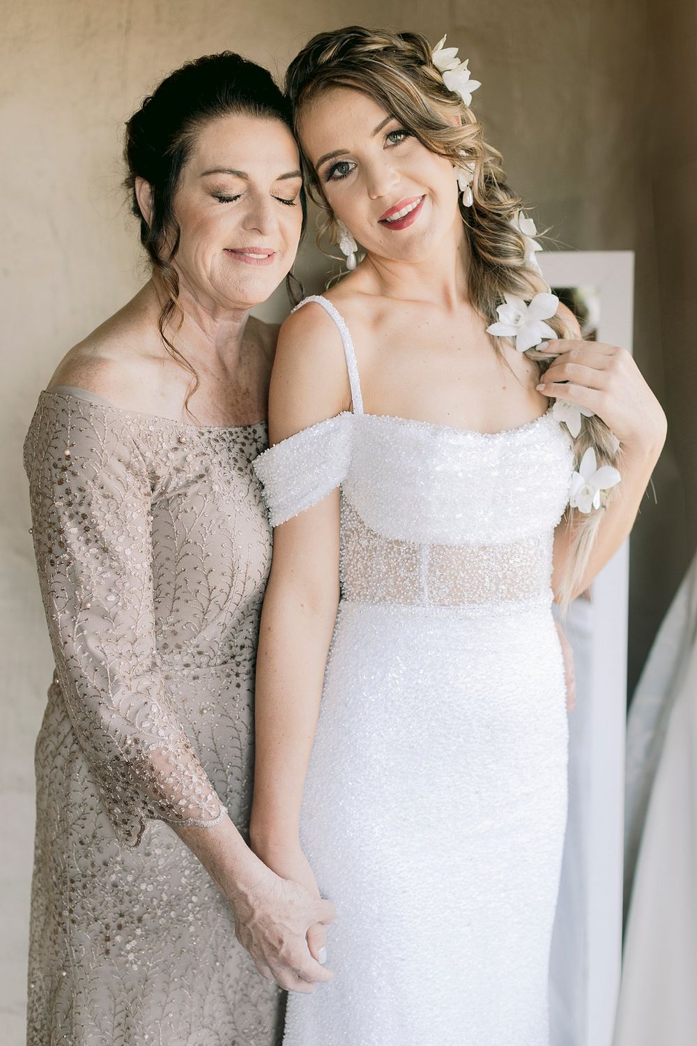 Smiling bride in white sequined gown with floral hair accessories embracing older woman in silver lace dress.