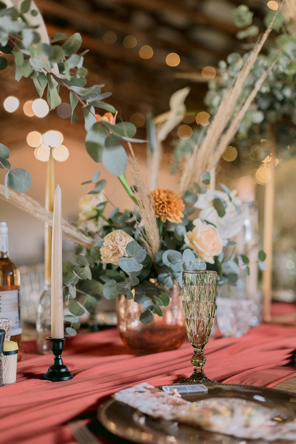 Elegant wedding table with red linen, taper candles, eucalyptus centerpiece, and green goblet with warm bokeh lights.