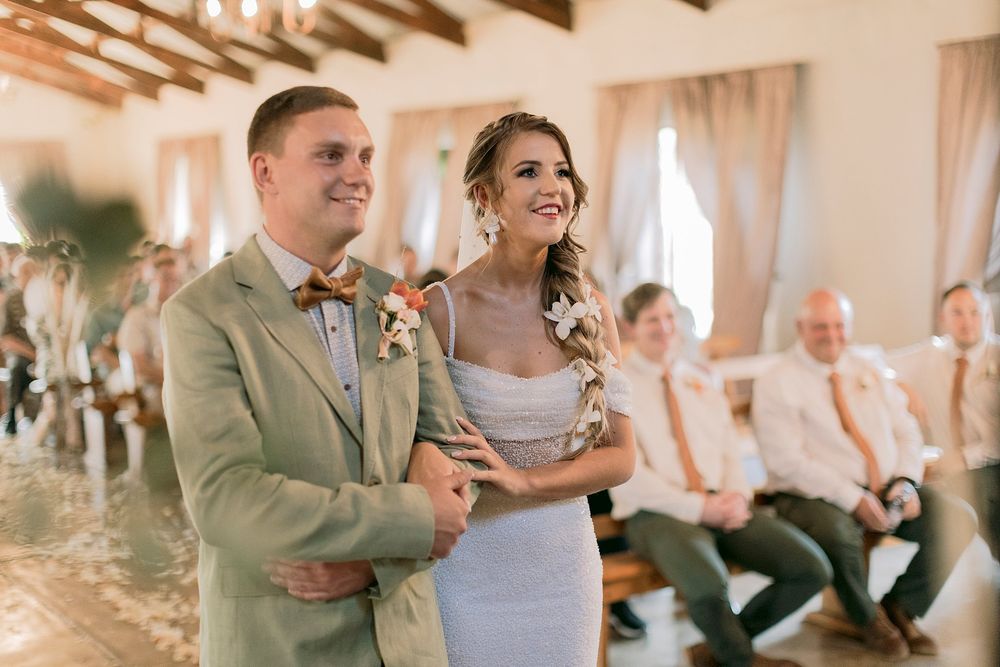 Smiling bride and groom in sage suit stand together during wedding ceremony in a sunlit rustic venue.