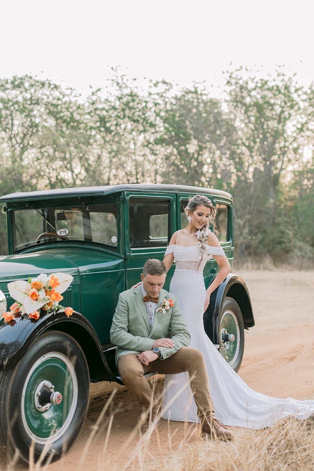 Bride in elegant white gown stands beside groom seated against a vintage green car on sandy terrain.