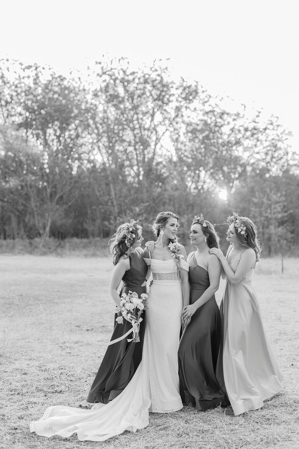 Black and white photo of a bride and three bridesmaids in elegant gowns sharing a joyful moment outdoors.