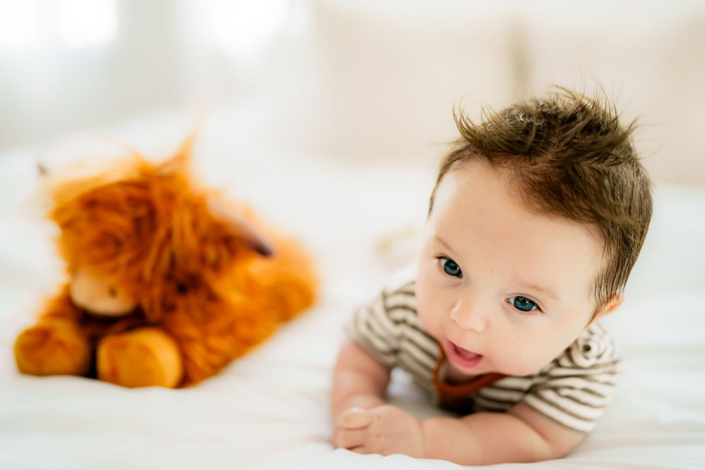 Baby in striped shirt lying on white bedding next to orange plush toy cat.