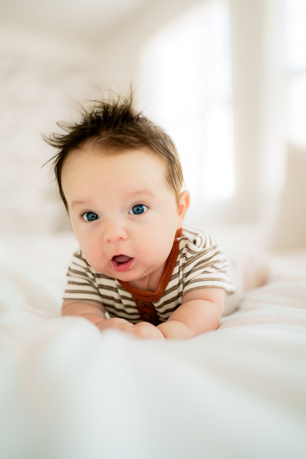 Baby in striped onesie lying on white bedding with bright eyes and tousled hair.