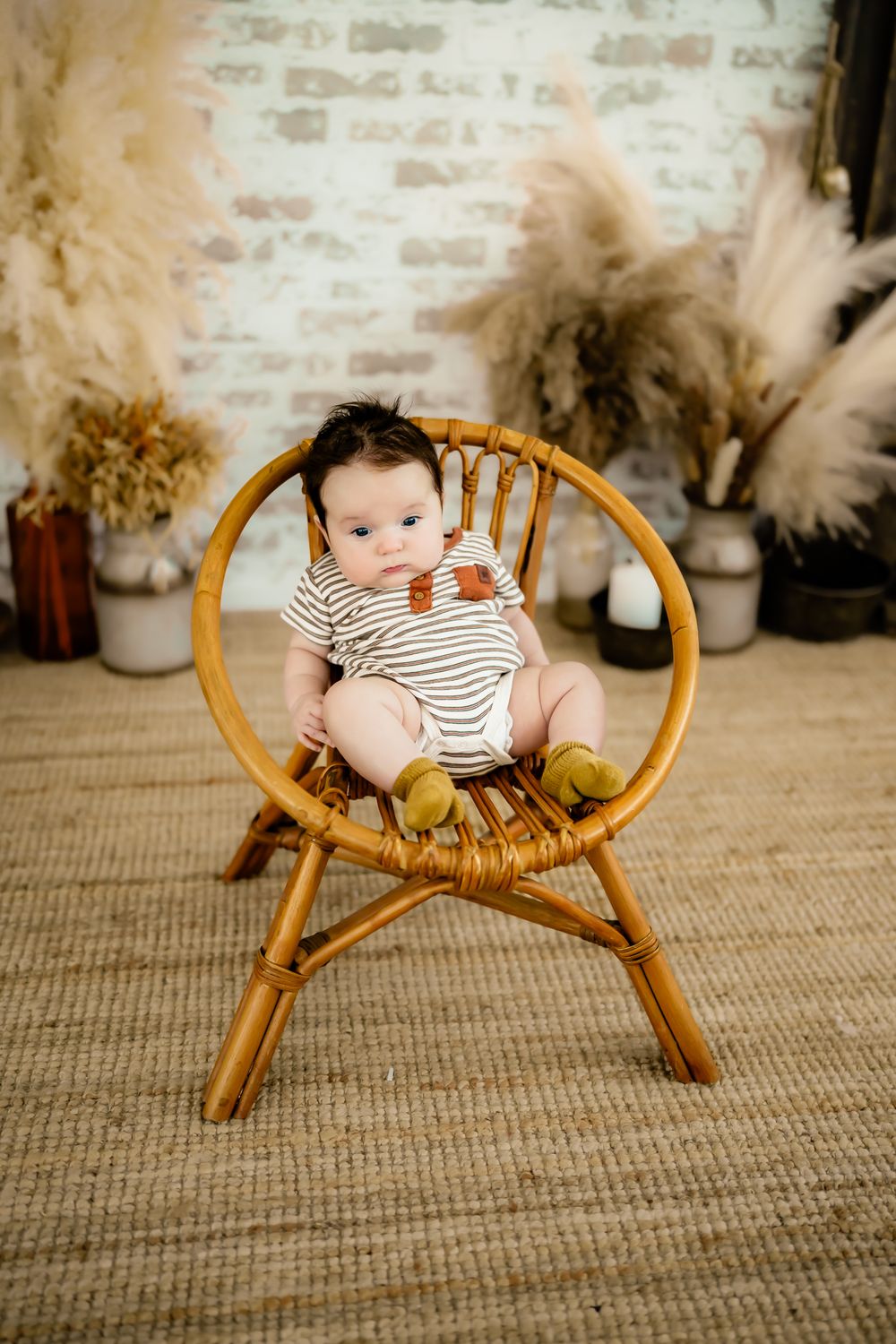 Baby sitting in vintage rattan chair with striped outfit against brick wall and pampas grass decor.