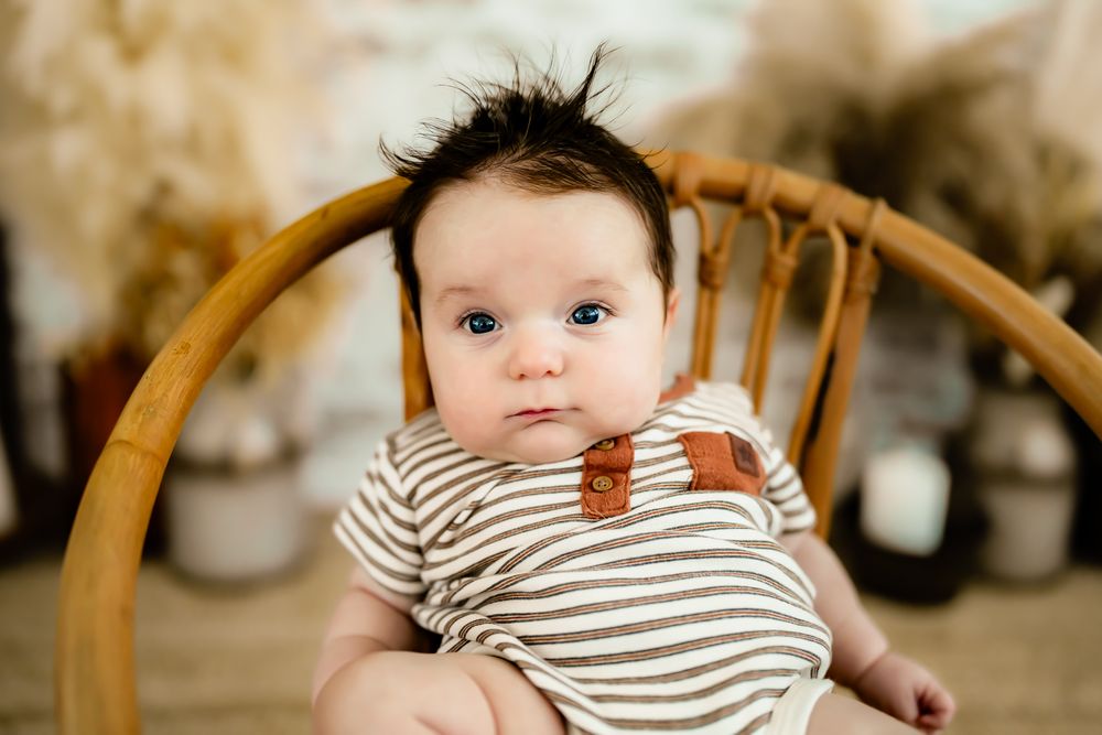 Baby in striped outfit sitting in wooden chair with spiky hair and bright eyes.