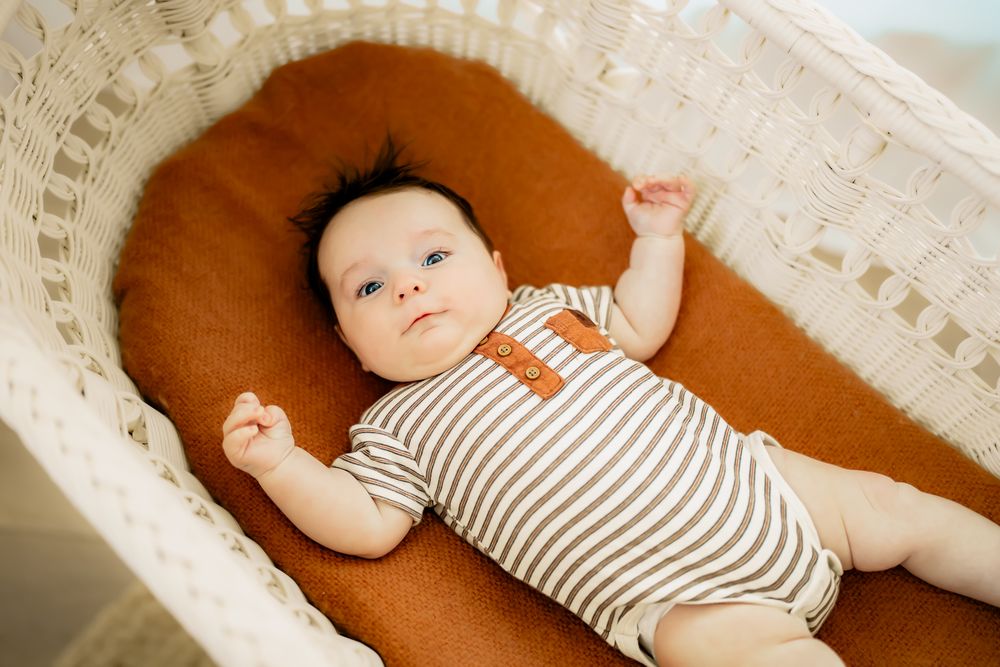 Baby wearing striped onesie lies contentedly on brown cushion in wicker bassinet.