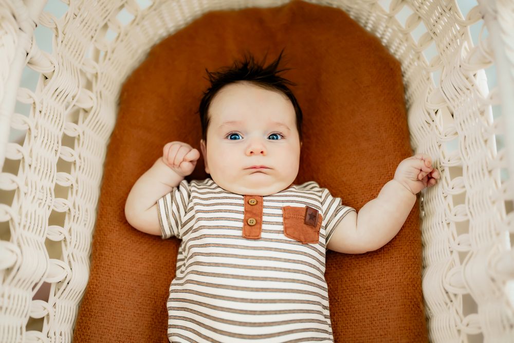 Baby in striped onesie lying on rust-colored cushion with arms outstretched.
