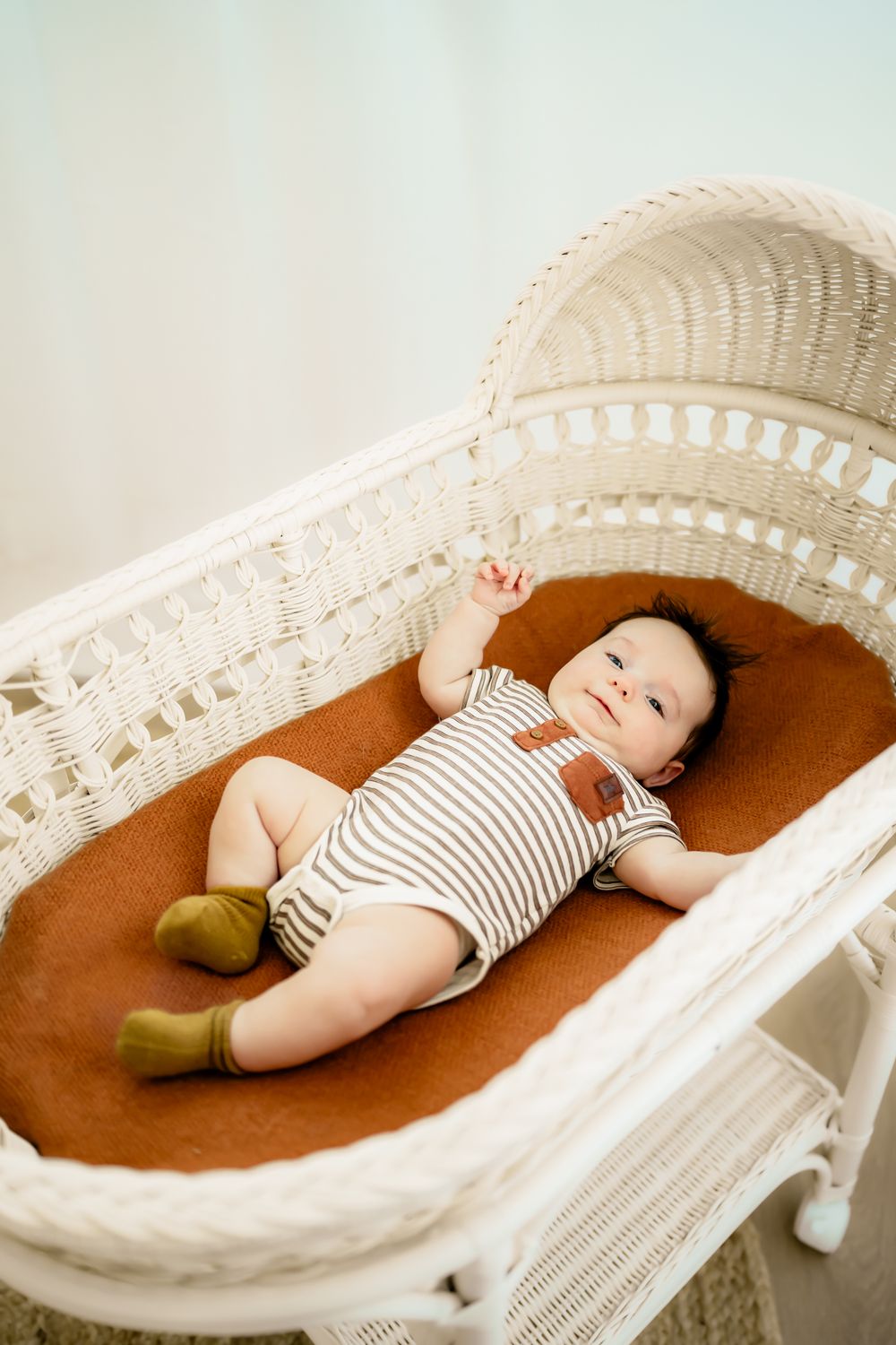 Baby wearing striped onesie lying in white wicker bassinet on rust-colored cushion.
