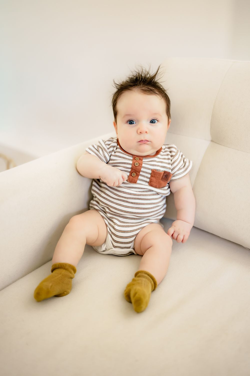 Baby wearing striped onesie with brown accents and mustard colored socks sits on white couch.