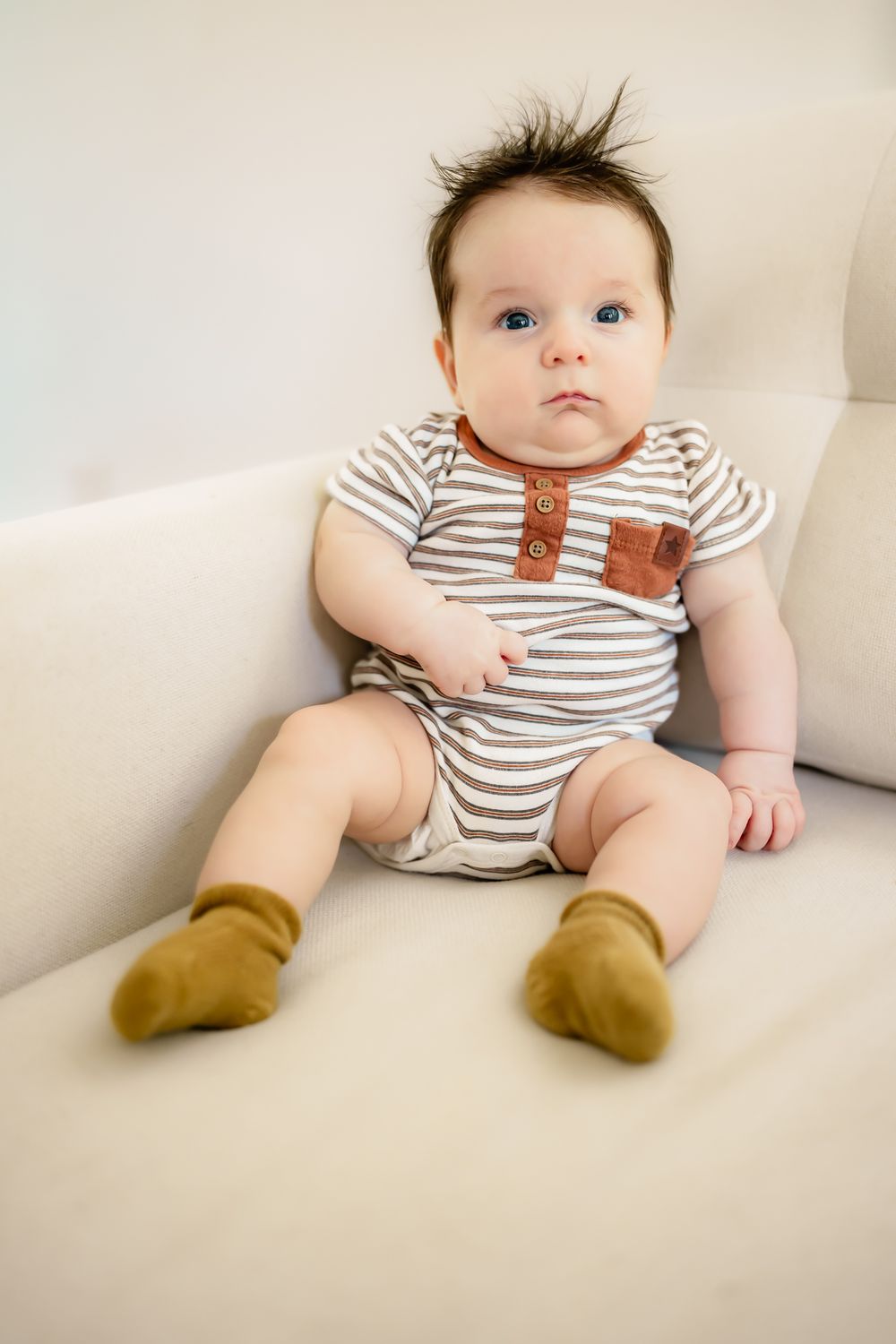 Baby wearing striped onesie with brown accents and mustard socks sits on cream couch.