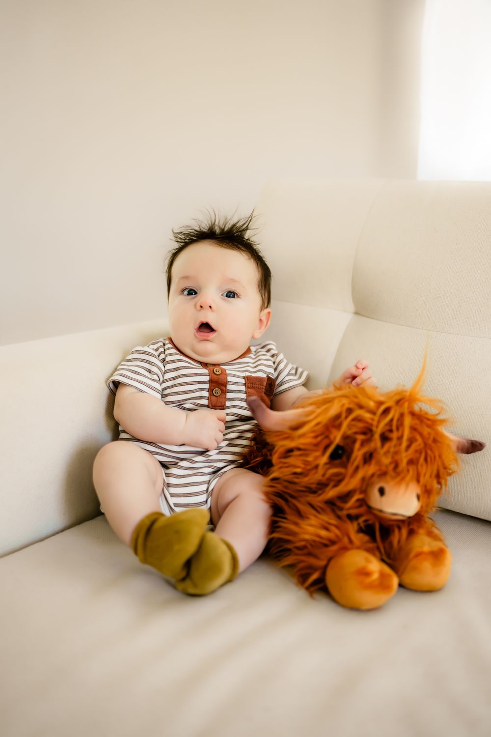 Small baby in striped onesie sitting on white couch with orange plush toy.