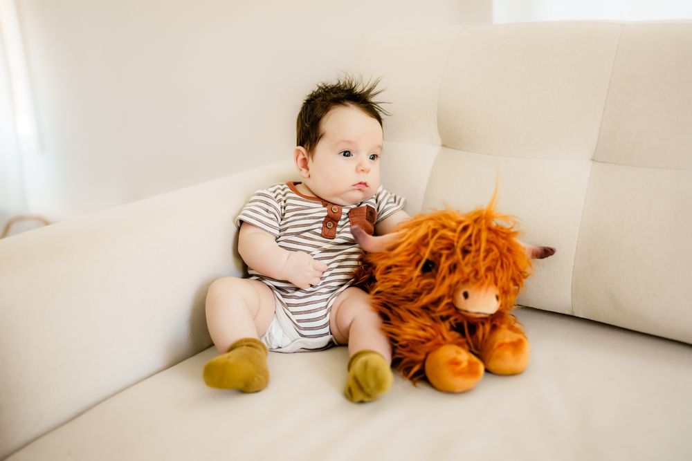 A child in a striped onesie sits on a white couch next to a large plush orange lion toy.