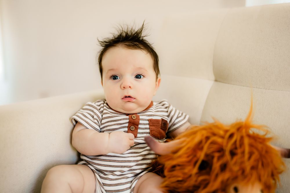 Baby wearing striped onesie sits on white couch next to orange plush toy.