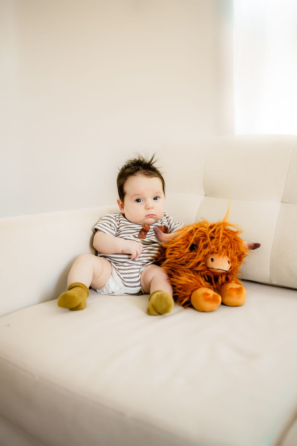 Small child in striped onesie sits on white couch with orange plush lion toy.