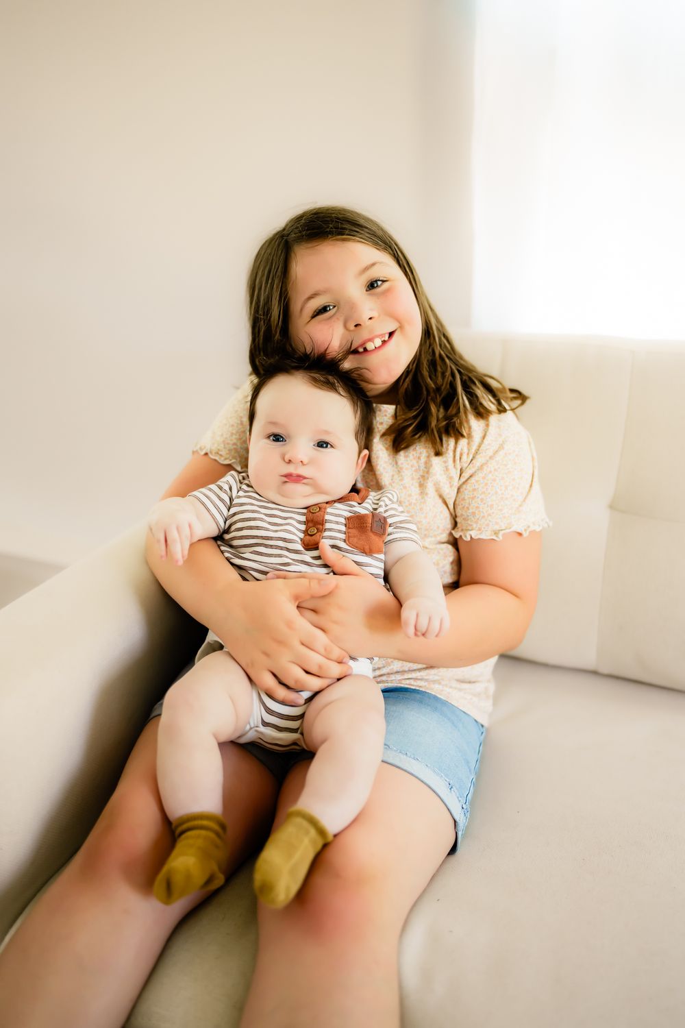 Two children sitting together on a sofa in a bright room, wearing casual clothes with the older one embracing the younger.