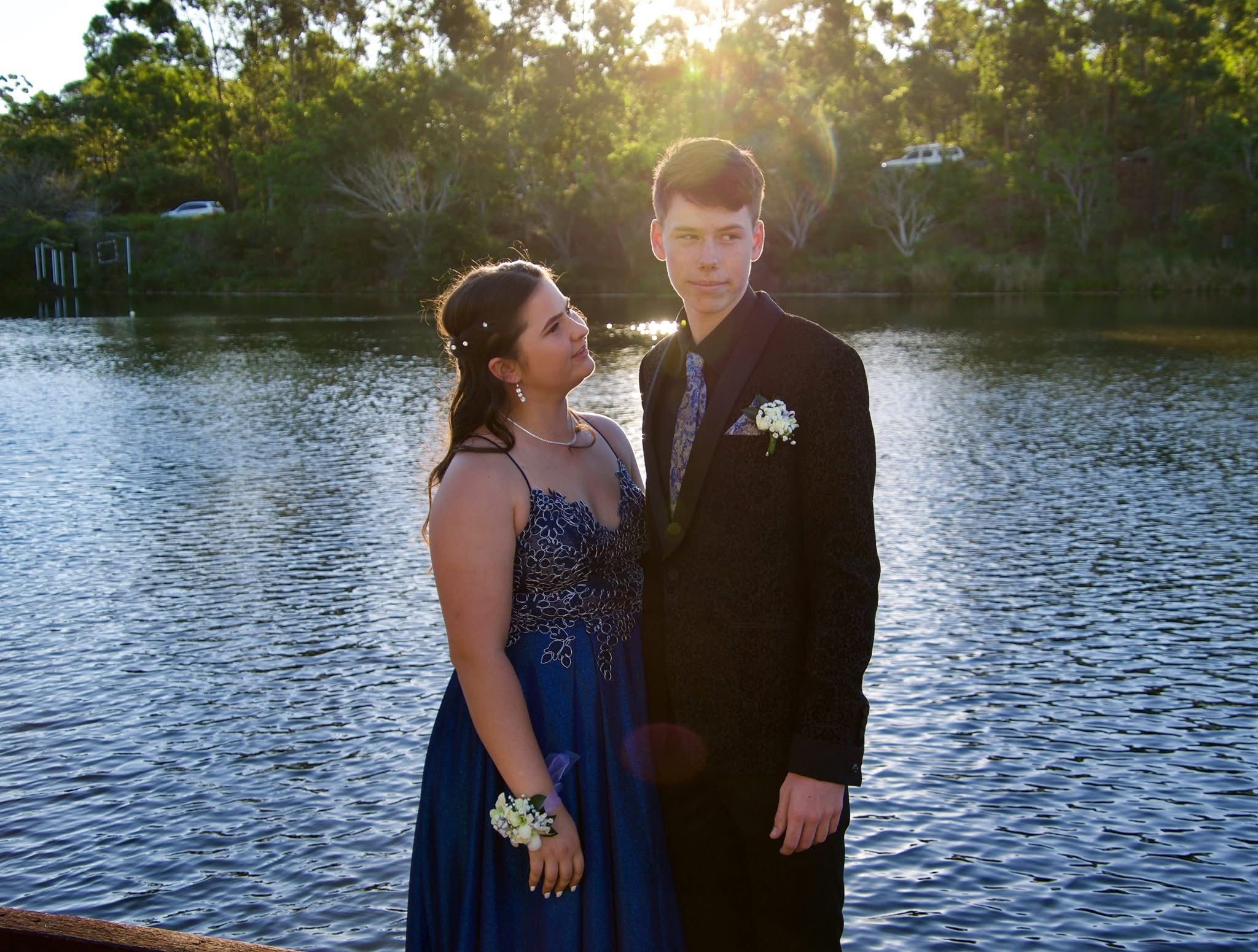 Couple in formal wear posing by a lake at sunset for prom photos.