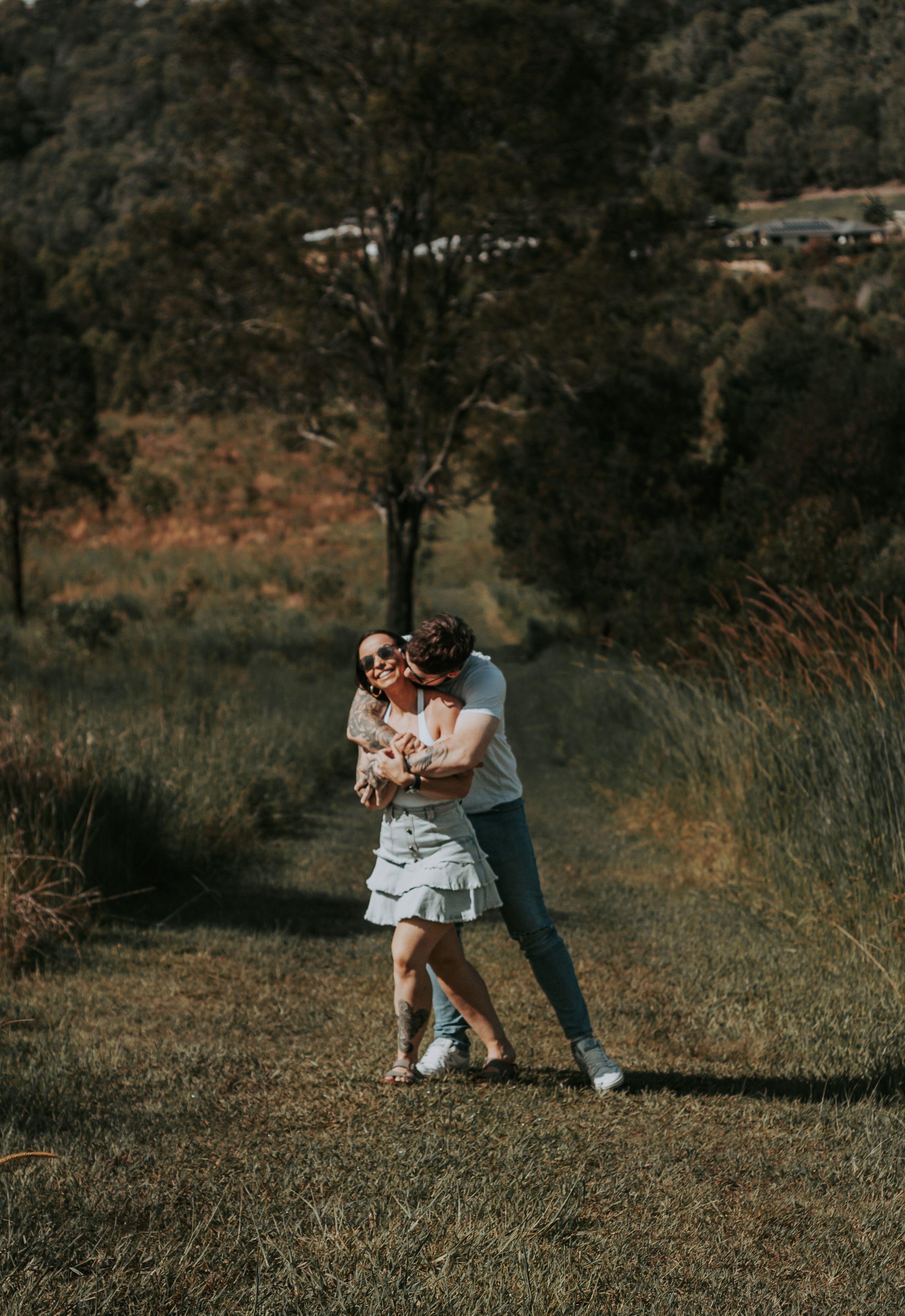 A couple shares a tender embrace in a sunlit field with mountains in the background.