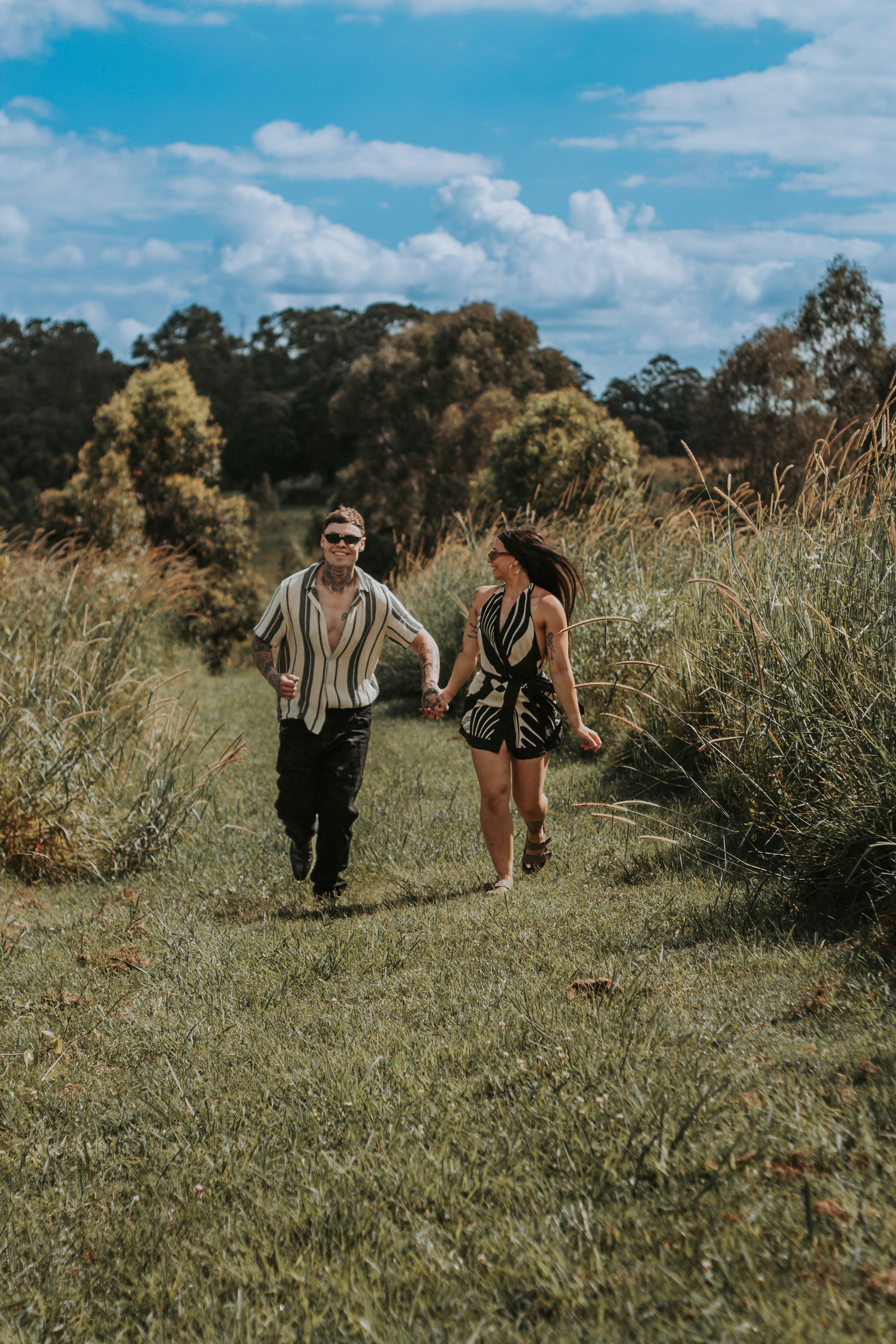A couple walks hand in hand through a grassy field on a sunny day.