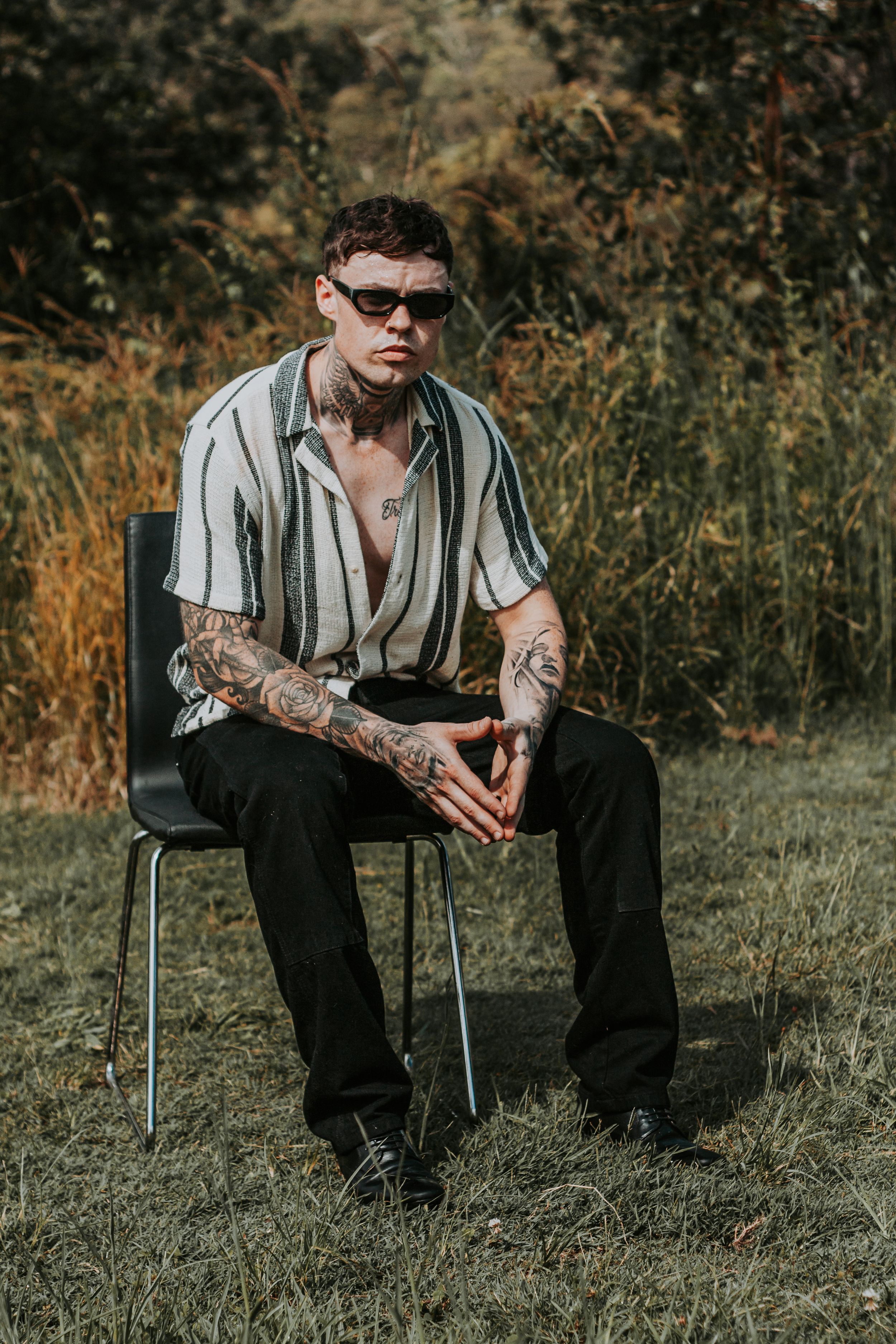 A tattooed figure in sunglasses and striped shirt sits on a chair in an outdoor setting.