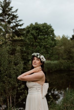 Bride in white dress and floral crown poses elegantly by a serene pond with evergreen trees.