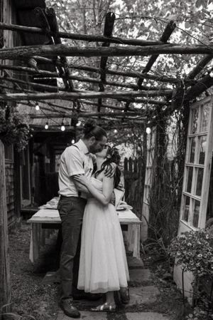 Black and white photo of couple sharing intimate moment under rustic wooden pergola.