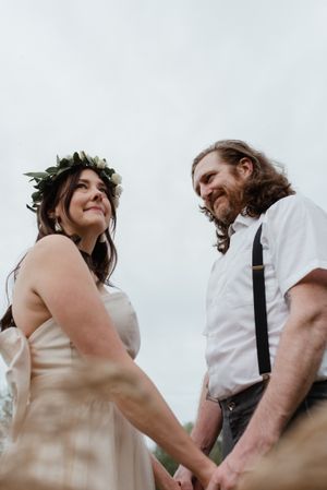 A tender wedding moment captured from below showing a couple holding hands, with the bride wearing a floral crown.