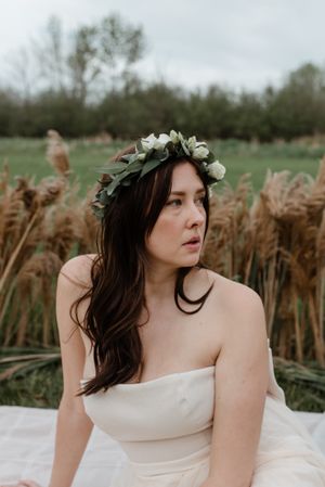 A delicate floral crown adorns long dark hair against a backdrop of tall wheat grass and cloudy skies.