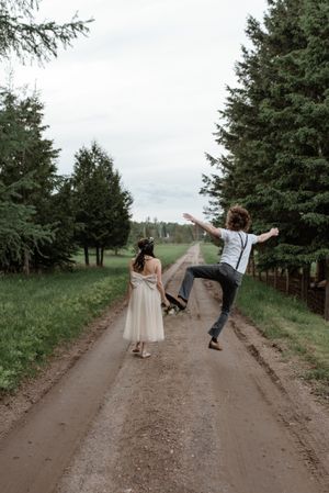 A joyful couple celebrates on a dirt road, with one person jumping while wearing suspenders.