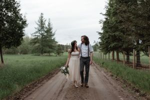 A couple in rustic wedding attire walks together down a tree-lined dirt path.