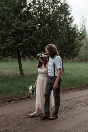A couple in vintage-inspired wedding attire stands together on a country road.