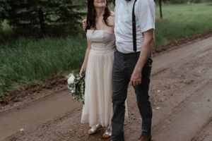 A couple in casual wedding attire stands together on a rustic dirt road.