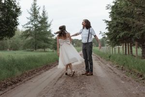 A couple dances playfully together on a dirt road surrounded by green trees.