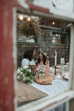 A cozy outdoor dinner setting viewed through a window with hanging lights and floral arrangements.