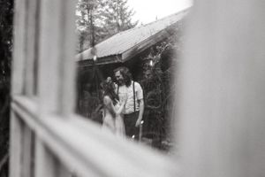 A romantic black and white photo of a couple standing near a rustic cabin in the woods.