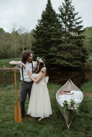 Romantic couple poses with a wooden canoe and paddle by a serene pond surrounded by evergreens.