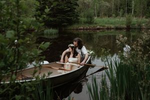 A romantic couple sits in a wooden rowboat on a serene pond surrounded by cattails and greenery.