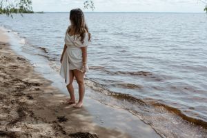 Woman in flowing white dress walking barefoot along shoreline.
