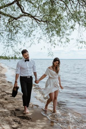 Couple walking hand in hand on beach shore under overhanging tree branches.