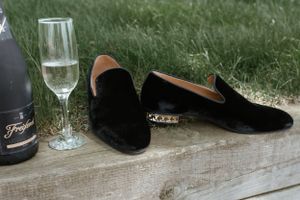 Black velvet loafers and champagne glass arranged on rustic wooden surface in outdoor setting.