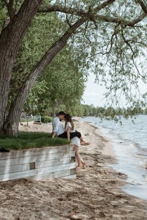 Couple sitting on wooden fence by lake shore under overhanging tree branches.